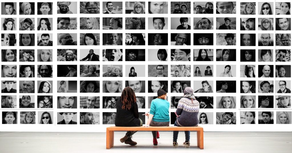 3 pessoas sentadas em um banco olhando um mural de fotos com várias pessoas de nacionalidades diversas