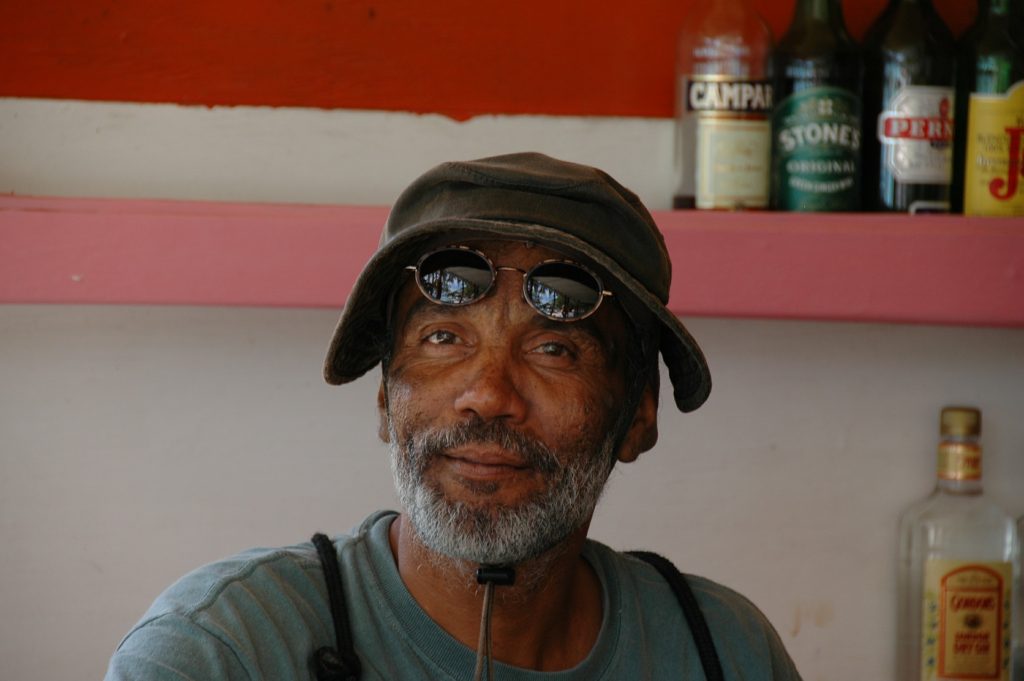 Homem negro, de barba grisalha, dono de um bar, usando chapéu, óculos na testa, com um sorriso de realizado