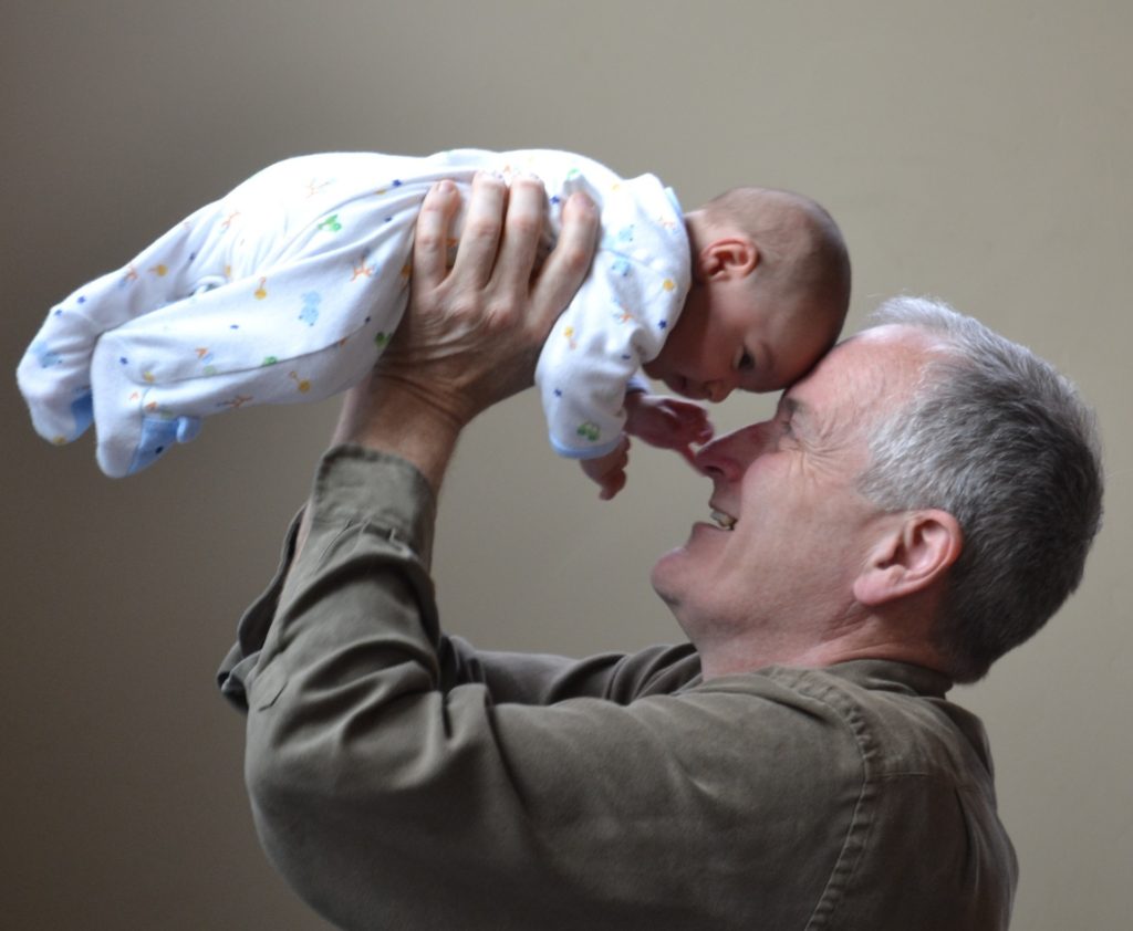 Homem grisalho, branco, feliz, levantando um bebê sobre a cabeça