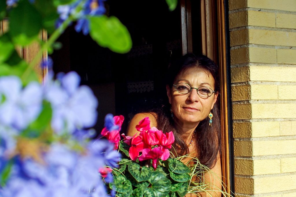 Mulher morena, de cabelos compridos castanhos, com óculos de grau, posando na janela com uma flor rosa em destaque